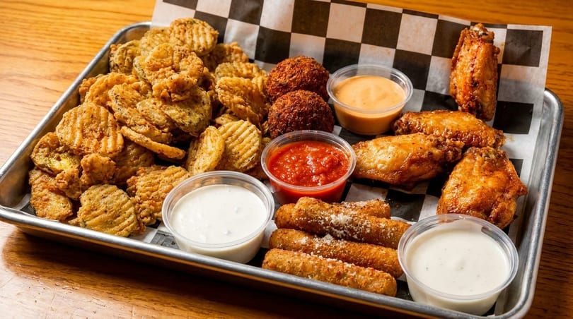 A metal tray filled with fried pickles, hush puppies, mozzarella sticks, chicken wings, and dipping sauces.